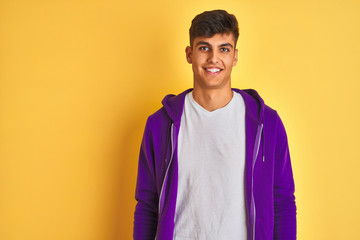 Young indian man wearing purple sweatshirt standing over isolated yellow background with a happy and cool smile on face. Lucky person.