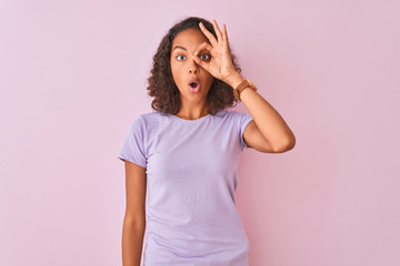 Young brazilian woman wearing t-shirt standing over isolated pink background doing ok gesture shocked with surprised face, eye looking through fingers. Unbelieving expression.