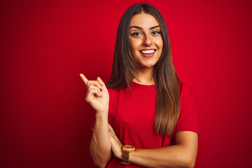 Young beautiful woman wearing t-shirt standing over isolated red background with a big smile on face, pointing with hand and finger to the side looking at the camera.