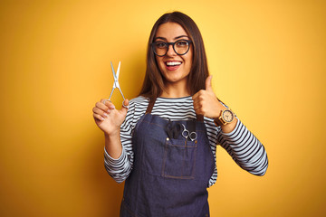 Young beautiful hairdresser woman holding scissors standing over isolated yellow background happy with big smile doing ok sign, thumb up with fingers, excellent sign