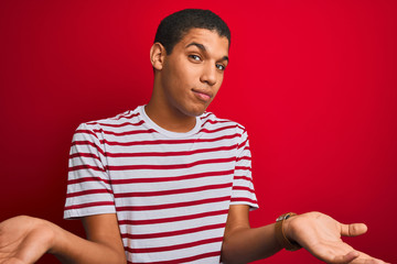Young handsome arab man wearing striped t-shirt over isolated red background clueless and confused with open arms, no idea concept.