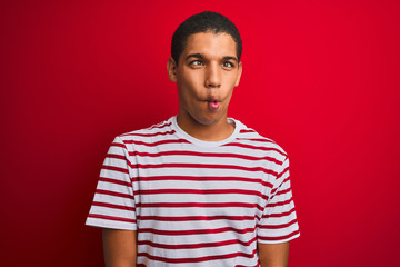 Young handsome arab man wearing striped t-shirt over isolated red background making fish face with lips, crazy and comical gesture. Funny expression.