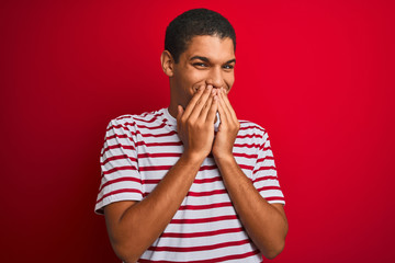 Young handsome arab man wearing striped t-shirt over isolated red background laughing and embarrassed giggle covering mouth with hands, gossip and scandal concept