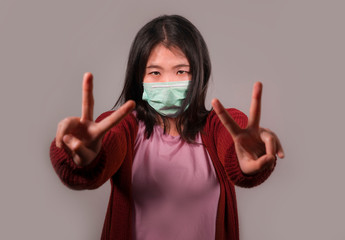 Young beautiful pacifist Asian Chinese woman in face mask doing peace fingers symbol standing for...