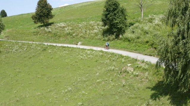 Aerial pan of two people riding bicycles on a country road driving towards a small chapel in remote nature in Prevalje, Slovenia