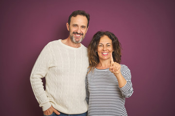 Beautiful middle age couple wearing winter sweater over isolated purple background showing and pointing up with finger number one while smiling confident and happy.