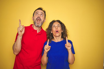 Beautiful middle age couple together standing over isolated yellow background amazed and surprised looking up and pointing with fingers and raised arms.