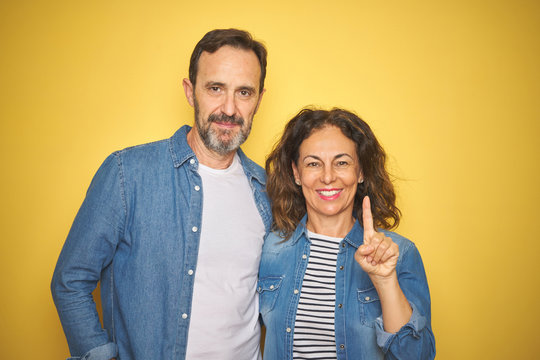 Beautiful middle age couple together wearing denim shirt over isolated yellow background showing and pointing up with finger number one while smiling confident and happy.