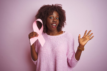 Young african afro woman holding cancer ribbon standing over isolated pink background very happy and excited, winner expression celebrating victory screaming with big smile and raised hands