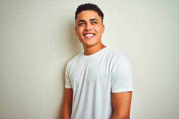 Young brazilian man wearing t-shirt standing over isolated white background looking away to side with smile on face, natural expression. Laughing confident.