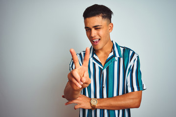 Young brazilian man wearing summer striped shirt standing over isolated white background smiling with happy face winking at the camera doing victory sign. Number two.