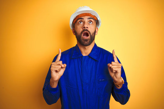 Handsome indian worker man wearing uniform and helmet over isolated yellow background amazed and surprised looking up and pointing with fingers and raised arms.