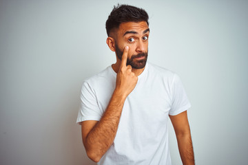 Young indian man wearing t-shirt standing over isolated white background Pointing to the eye watching you gesture, suspicious expression