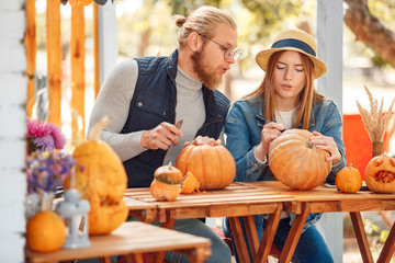 Halloween Preparaton Concept. Young couple sitting at table outdoors making jack-o'-lantern drawing...
