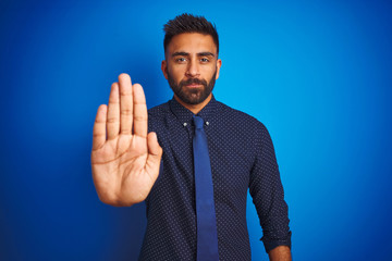 Young indian businessman wearing elegant shirt and tie standing over isolated blue background doing stop sing with palm of the hand. Warning expression with negative and serious gesture on the face.