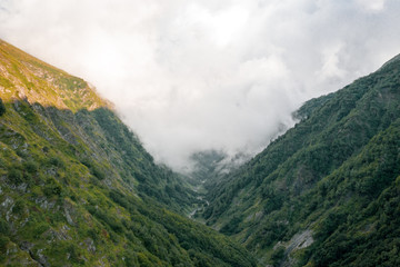 Fototapeta premium AERIAL 4K; drone flying forward between mountain ranges in deep dark river valley, low clouds; last evening sunbeams on steep overgreen slope; majestic foggy gorge, shadows of peaks, Caucasus, Russia