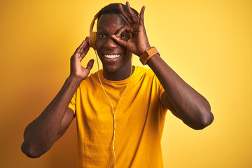 African american man listening to music using headphones over isolated yellow background with happy face smiling doing ok sign with hand on eye looking through fingers