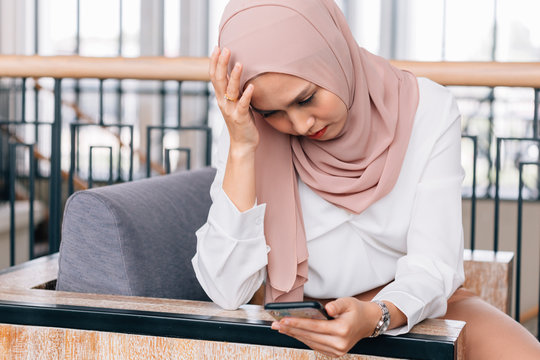 Sad Asian Female In Hijab Touching Forehead And Browsing Smartphone While Having Problem In Cafe