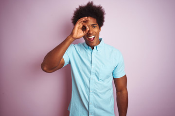 Young american man with afro hair wearing blue shirt standing over isolated pink background doing ok gesture with hand smiling, eye looking through fingers with happy face.