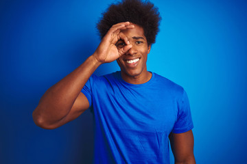 African american man with afro hair wearing t-shirt standing over isolated blue background doing ok gesture with hand smiling, eye looking through fingers with happy face.