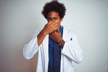 Fototapeta na wymiar Young african american doctor man wearing coat standing over isolated white background smelling something stinky and disgusting, intolerable smell, holding breath with fingers on nose. Bad smells
