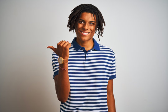 Afro man with dreadlocks wearing striped blue polo standing over isolated white background smiling with happy face looking and pointing to the side with thumb up.