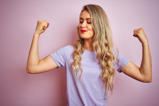 Young beautiful woman wearing purple t-shirt standing over pink isolated background showing arms muscles smiling proud. Fitness concept.