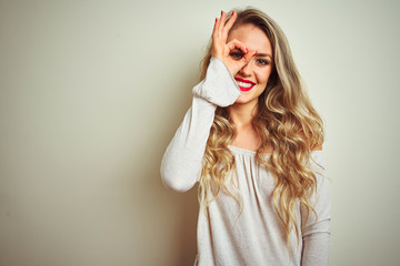 Young beautiful woman standing over white isolated background doing ok gesture with hand smiling, eye looking through fingers with happy face.