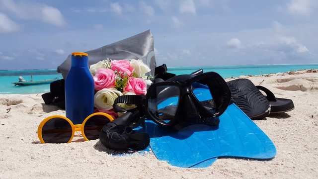 Stuff needed for a good day at the beach (snorkel, flipflops, sunglasses, sunblock and some roses) with boat at background with clear blue skies and clean waves on beautiful white sand -Closeup shot
