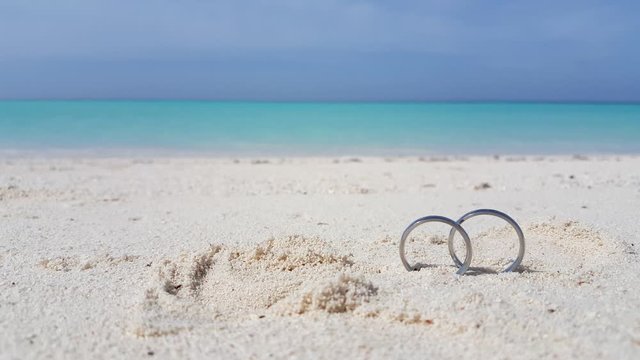 Wedding rings on the beautiful white sand beach with waves and clear blue sky at background in New Caledonia, France - Close up shot