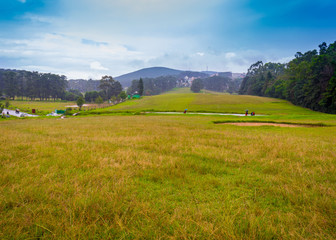 Golf course / meadow / grassland divided by road and surrounded by lush green forest, mountain and cloudy blue sky, Northeast, Shillong, Meghalaya, India.