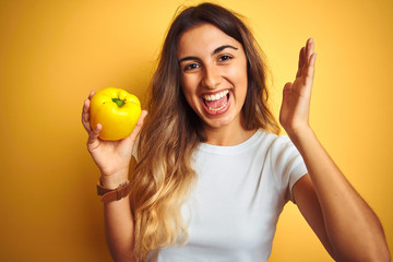 Young beautiful woman holding pepper over yellow isolated background very happy and excited, winner expression celebrating victory screaming with big smile and raised hands