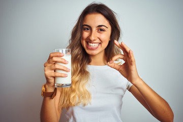 Young beautiful woman drinking a glass of milk over white isolated background doing ok sign with...