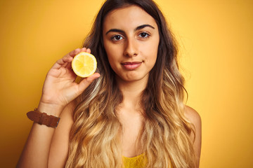 Young beautiful woman holding half lemon over yellow isolated background with a confident expression on smart face thinking serious