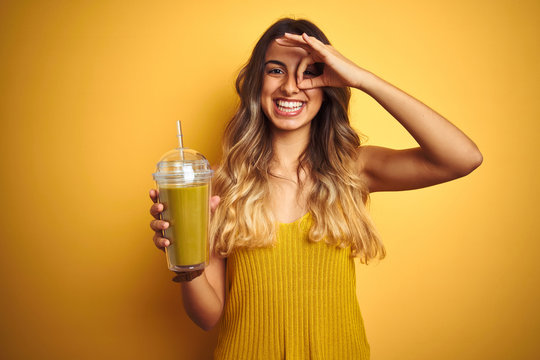 Young beautiful woman drinking detox green smoothy over yellow isolated background with happy face smiling doing ok sign with hand on eye looking through fingers