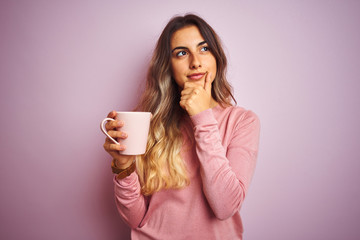 Young beautiful woman holding cup of coffee over pink isolated background serious face thinking...