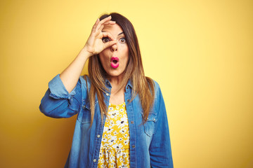 Young beautiful woman standing over yellow isolated background doing ok gesture shocked with surprised face, eye looking through fingers. Unbelieving expression.