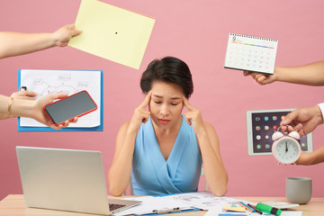 Overworked young employee refuses all things, frowns face in annoyance, sits at desktop with paper documents and notepad, isolated over pink background. Female workes bothered by many questions