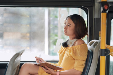 Beautiful young woman sitting in city bus and writing some notes in notebook.