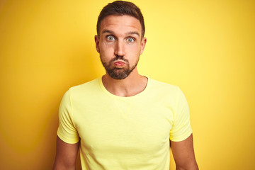 Young handsome man wearing casual yellow t-shirt over yellow isolated background puffing cheeks with funny face. Mouth inflated with air, crazy expression.
