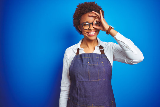 Young african american woman shop owner wearing business apron over blue background doing ok gesture with hand smiling, eye looking through fingers with happy face.