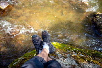 The feet of a resting man in white socks and black trekking sneakers lie on a moss-covered rock against a clear mountain river.