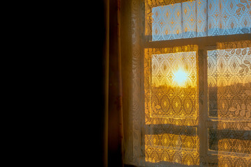 The bright yellow sunlight from the sunset over the horizon comes through the window in the village room with the white patterned curtain and the shadow of the frame into the dark part.