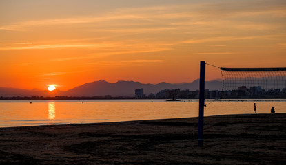 Beach volleyball court on a sandy beach by sea in evening. Amazing beautiful summer golden sunset over a bay of Roses, Catalunya, Spain. Sandy beach, calm sea on mountains and evening sky background.