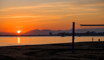 Fototapeta premium Beach volleyball court on a sandy beach by sea in evening. Amazing beautiful summer golden sunset over a bay of Roses, Catalunya, Spain. Sandy beach, calm sea on mountains and evening sky background.