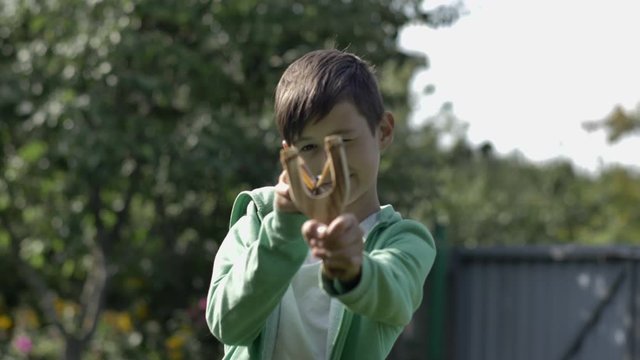 boy stands with a slingshot and aims at the camera in the village