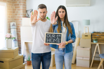 Fototapeta na wymiar Young beautiful couple holding blackboard with our first home text at new house with open hand doing stop sign with serious and confident expression, defense gesture