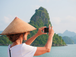 Woman with traditional hat using phone at Halong Bay, Vietnam. Tourist traveling on cruise among Ha...