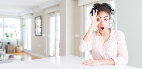 Wide angle of beautiful african american woman with afro hair doing ok gesture shocked with surprised face, eye looking through fingers. Unbelieving expression.