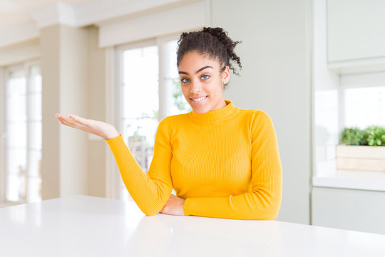 Beautiful african american woman with afro hair wearing a casual yellow sweater smiling cheerful presenting and pointing with palm of hand looking at the camera.